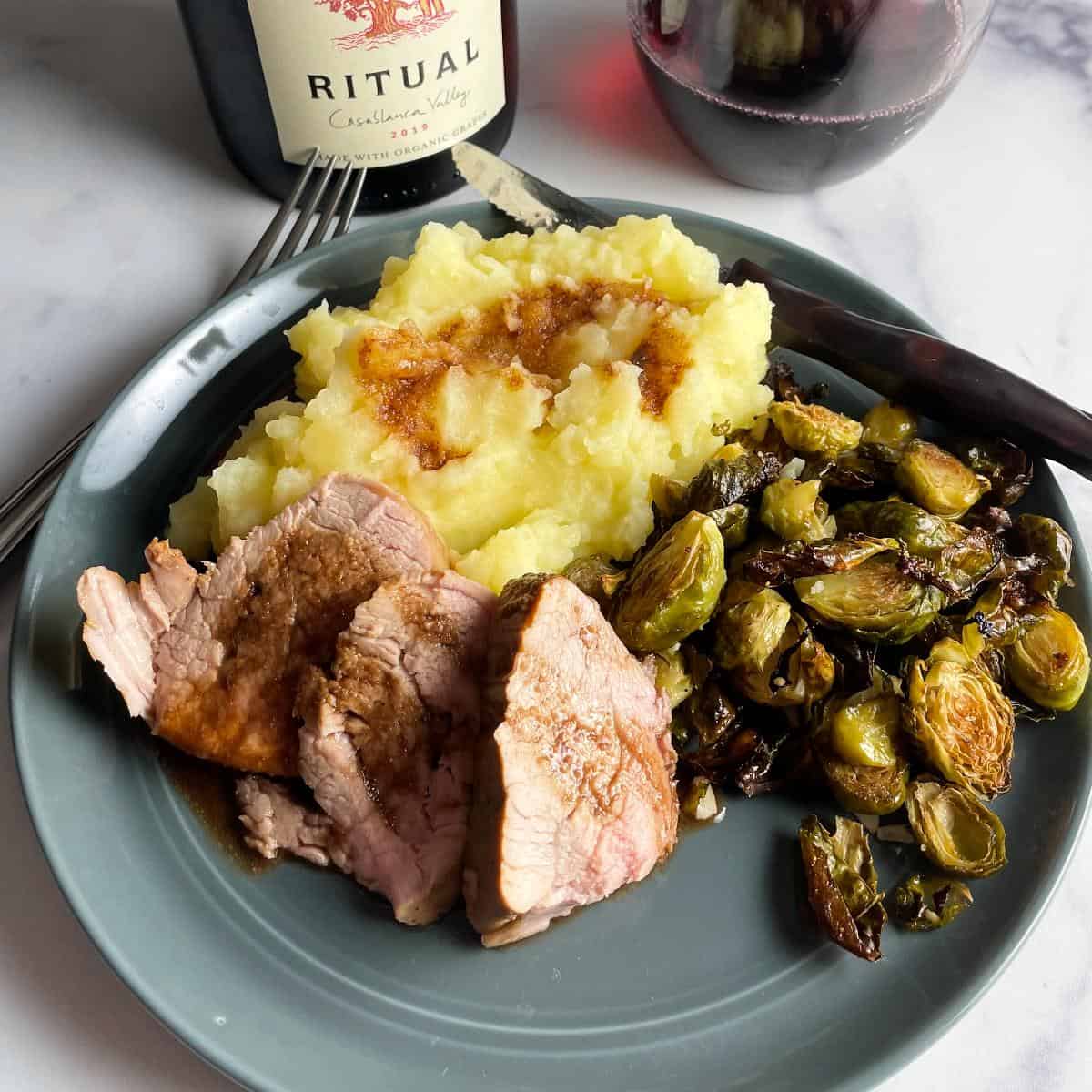 Slices of pork tenderloin plated with mashed potato and roasted Brussels sprouts, and some Ritual Pinot Noir red wine in the background.