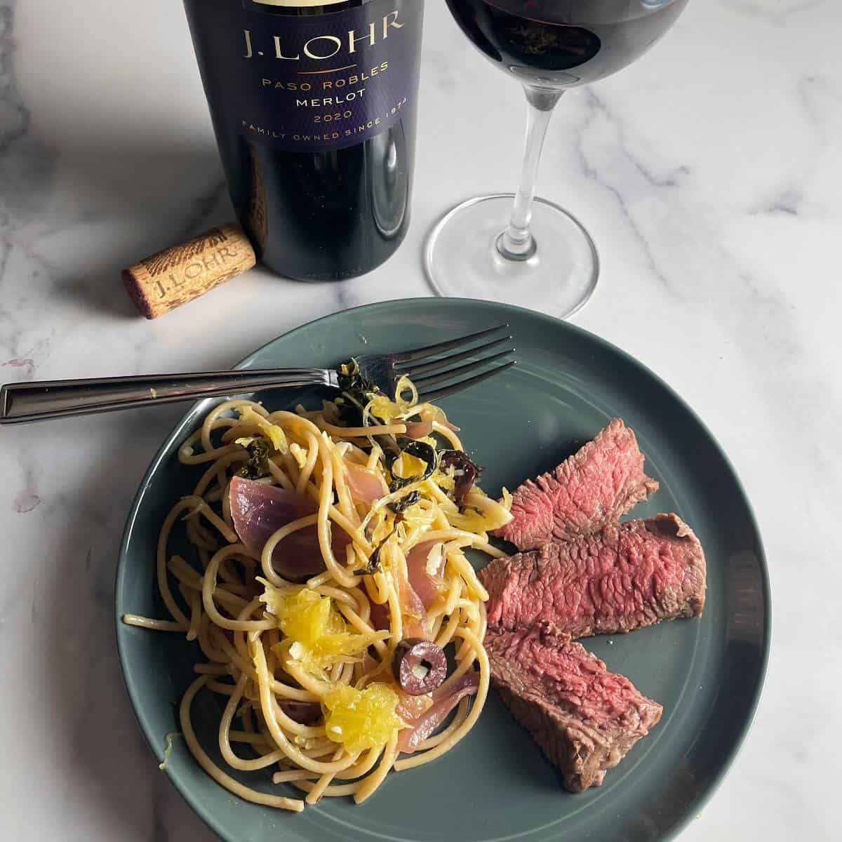 slices of steak served on a dark colored plate along with spaghetti and vegetables, and a glass of Merlot red wine.