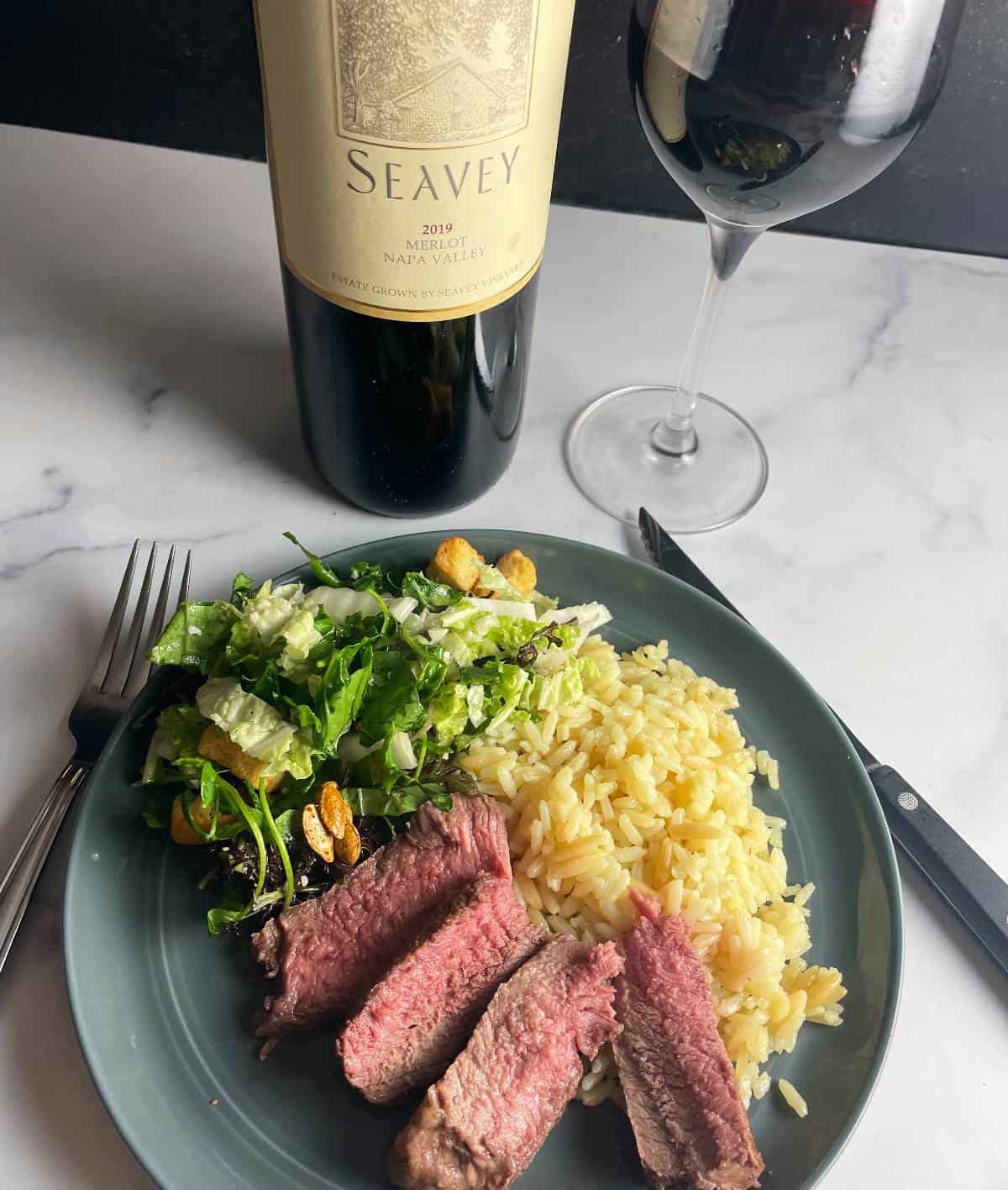 slices of steak served on a plate with rice and salad. Bottle of Merlot wine in the background.