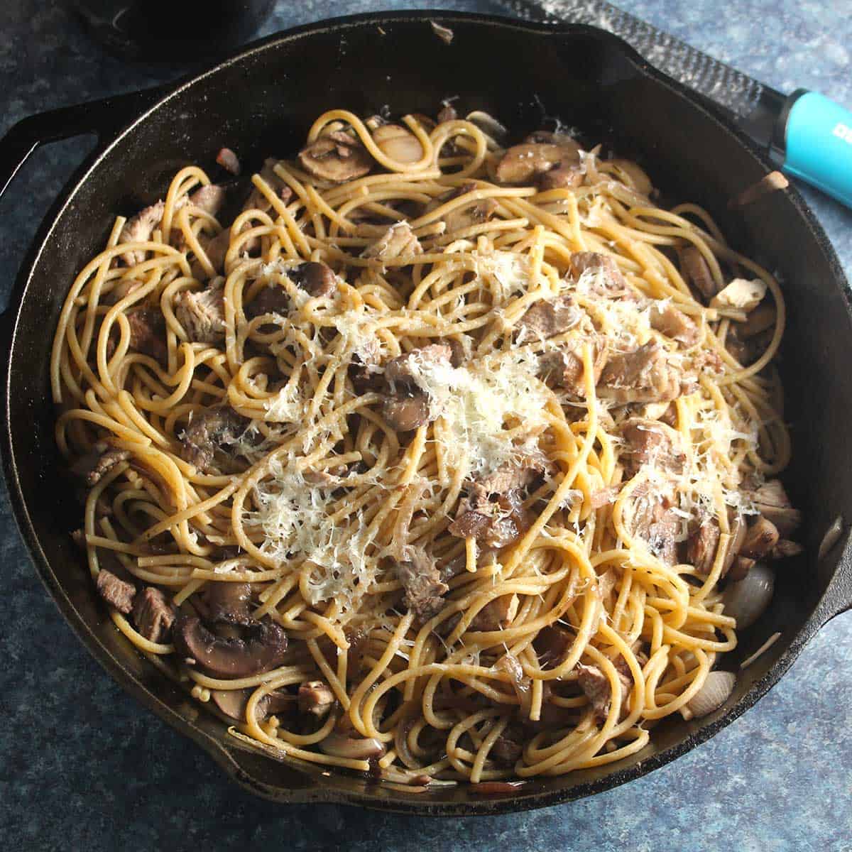 Spaghetti tossed with leftover turkey and mushrooms, topped with shredded cheese, in a black skillet. 