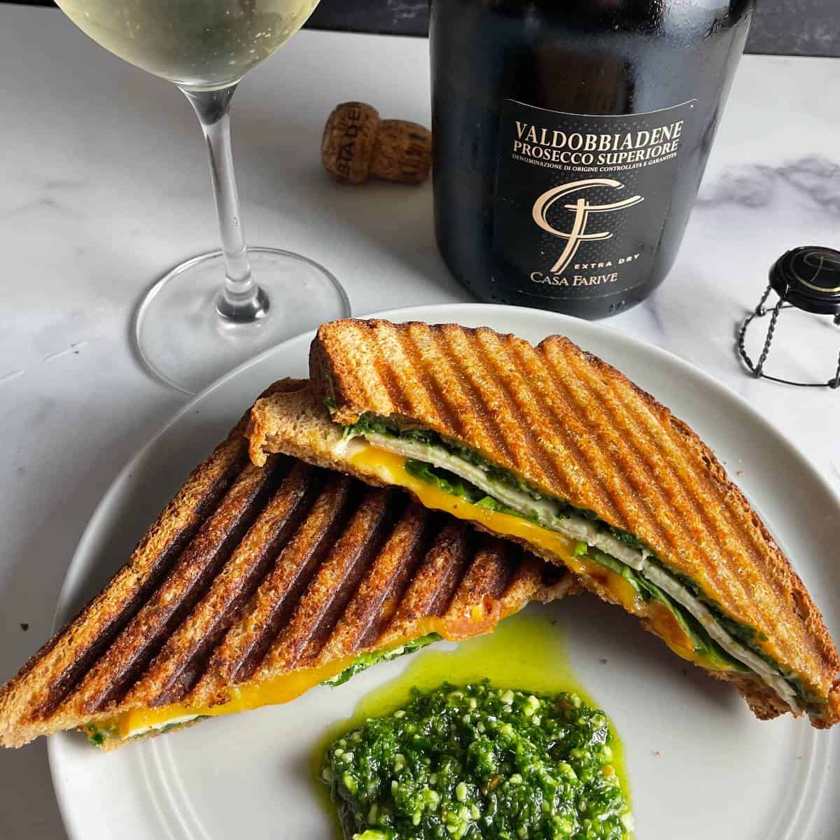 A turkey pesto panini, sliced in half and served along with an extra dollop of pesto. A bottle of Prosecco Superiore in the background.