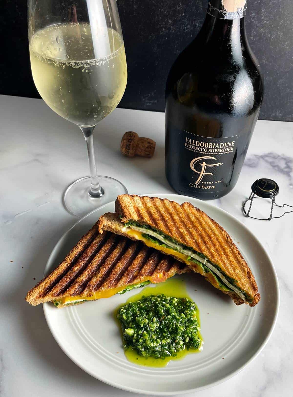Turkey Pesto Panini, sliced in half, and served with extra pesto. Shot from above. Bottle and glass of Prosecco Superiore in the background.