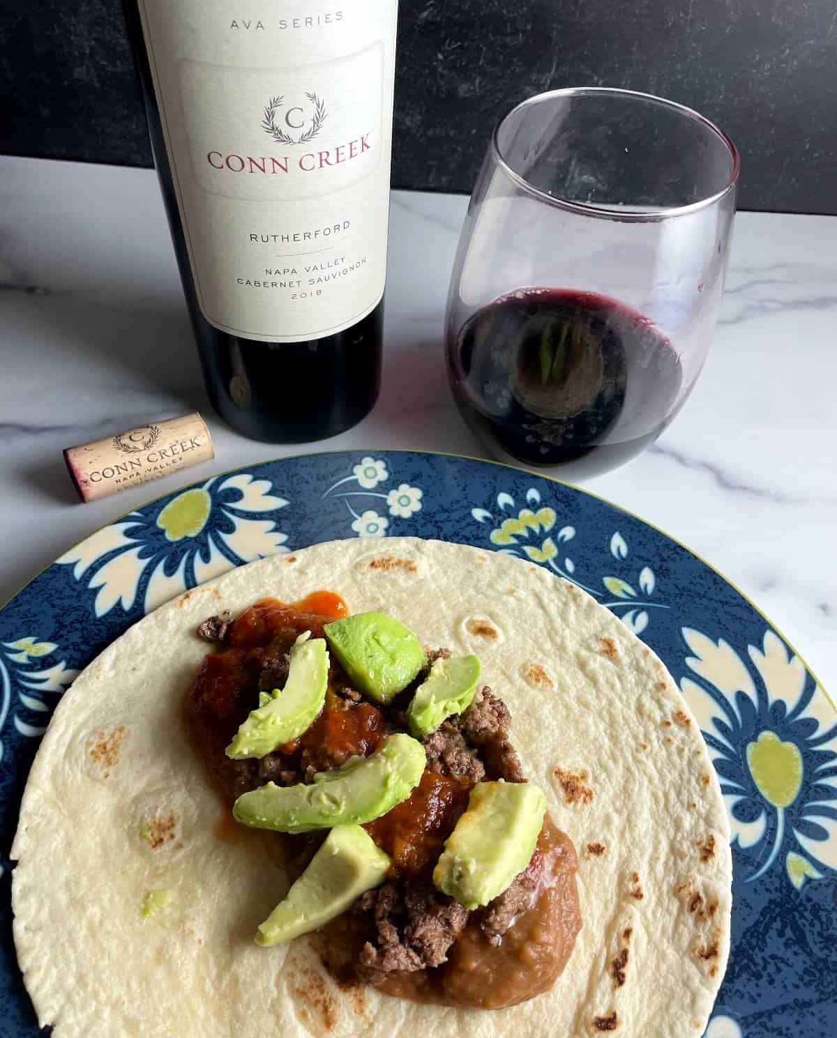 a beef and bean soft taco, topped with avocados, and served with a Cabernet Sauvignon red wine.
