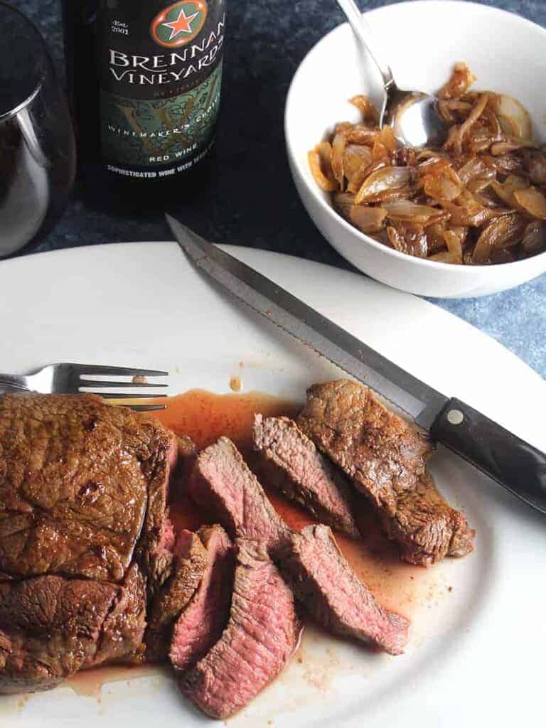slices of sirloin steak on a white platter, with an onions sauce and a bottle of red wine next to the platter.