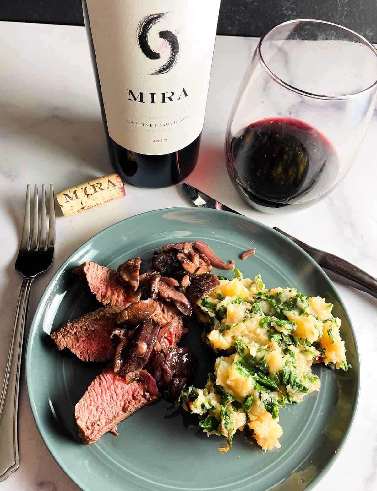 slices of steak topped with shiitake mushrooms, served with a side of Swiss chard potatoes. Bottle and glass of Mira Cabernet Sauvignon red wine in the background.