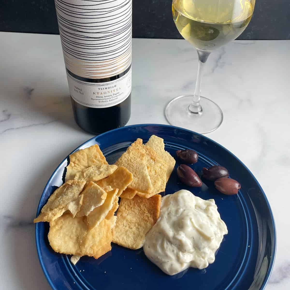 tzatziki dip on a blue plate with pita chips and kalamata olives, served with a Greek white wine.