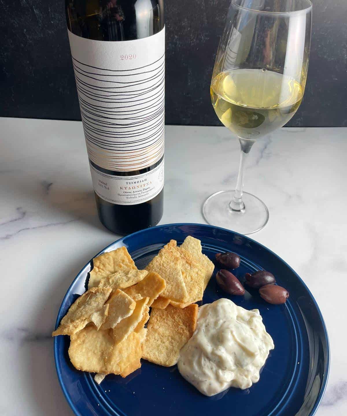 tzatziki dip on a blue plate with pita chips and kalamata olives, with a Greek white wine in the background.