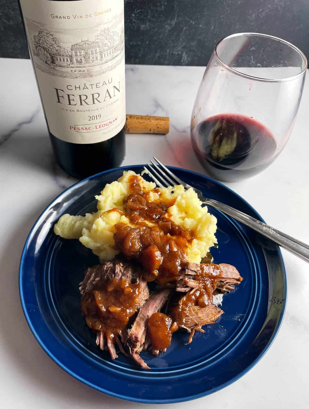 Braised brisket served with mashed potatoes on a blue plate, with Pessac-Léognan red wine in the background.