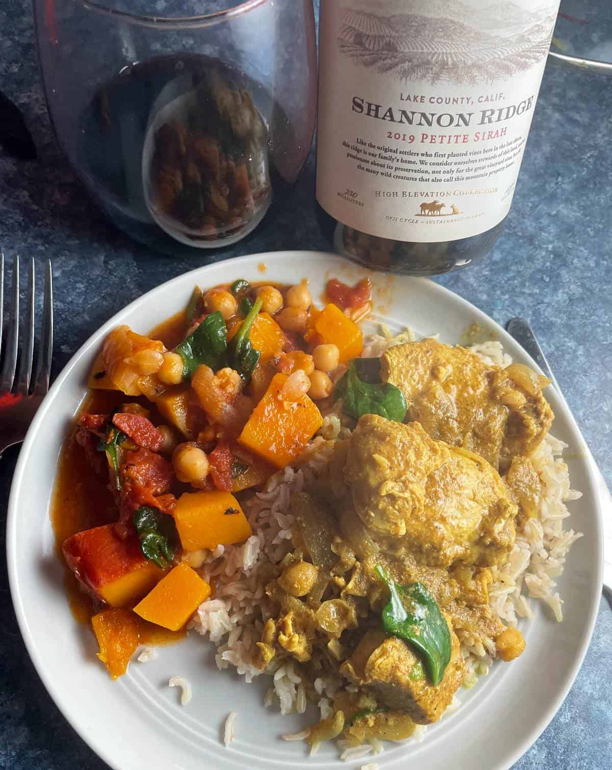 Indian butter chicken on a plate with butternut squash curry, and a bottle of red wine in the background.
