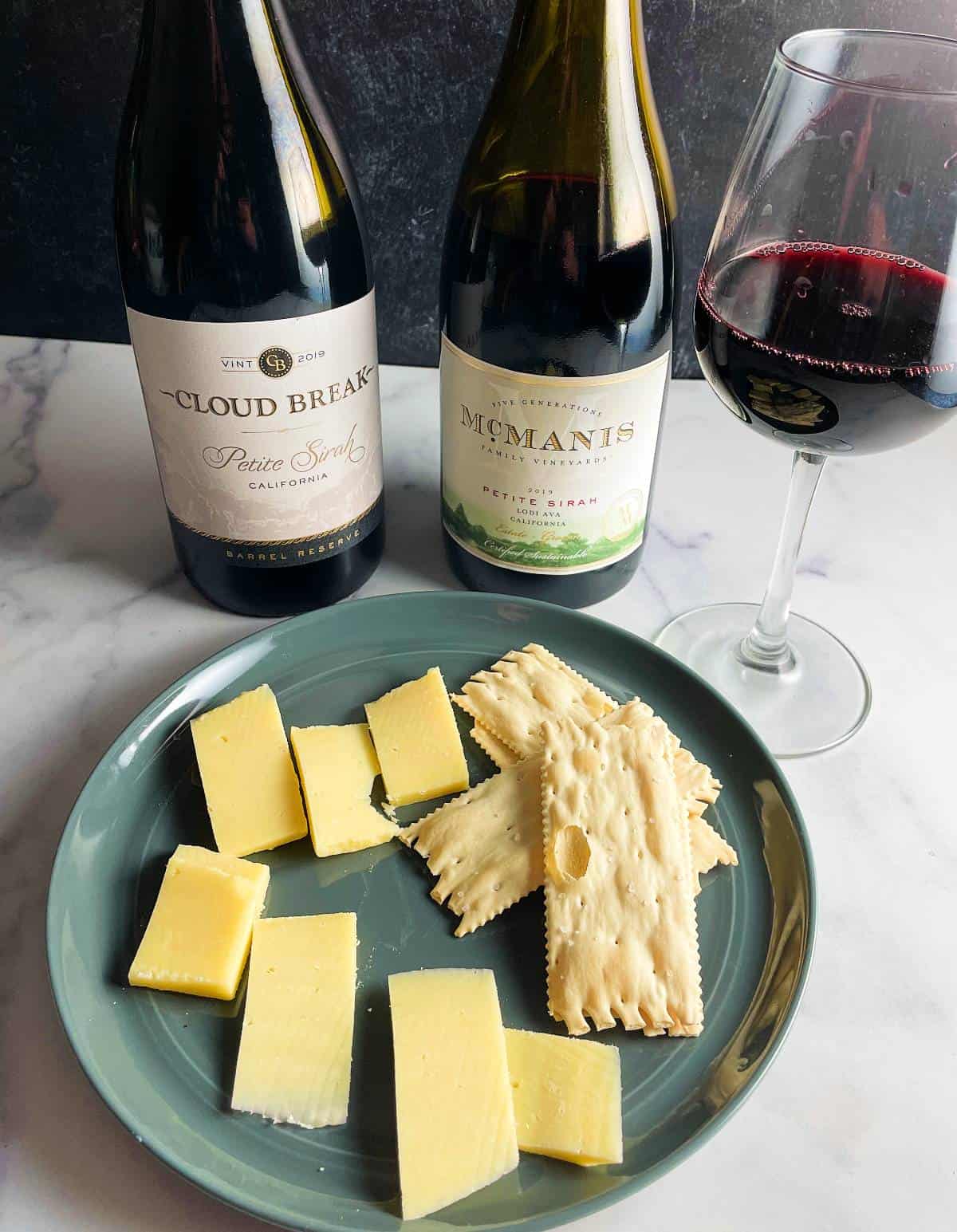 a turquoise plate with slices of cheddar cheese and crackers. Two bottles of Petite Sirah wine in the background.