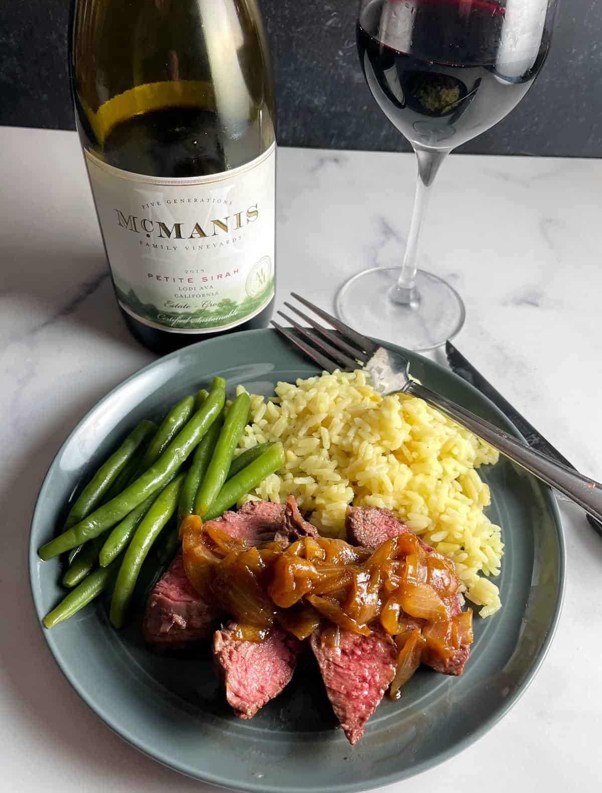 A dark gray plate with sliced steak topped with onion sauce, with sides of rice and green beans. Petite Sirah red wine in the background.