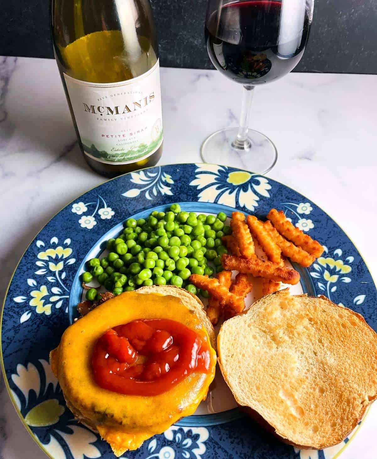 A veggie burger with cheese, topped with ketchup, with the top of the bun alongside the burger. Served with fries and peas, and Petite Sirah wine.