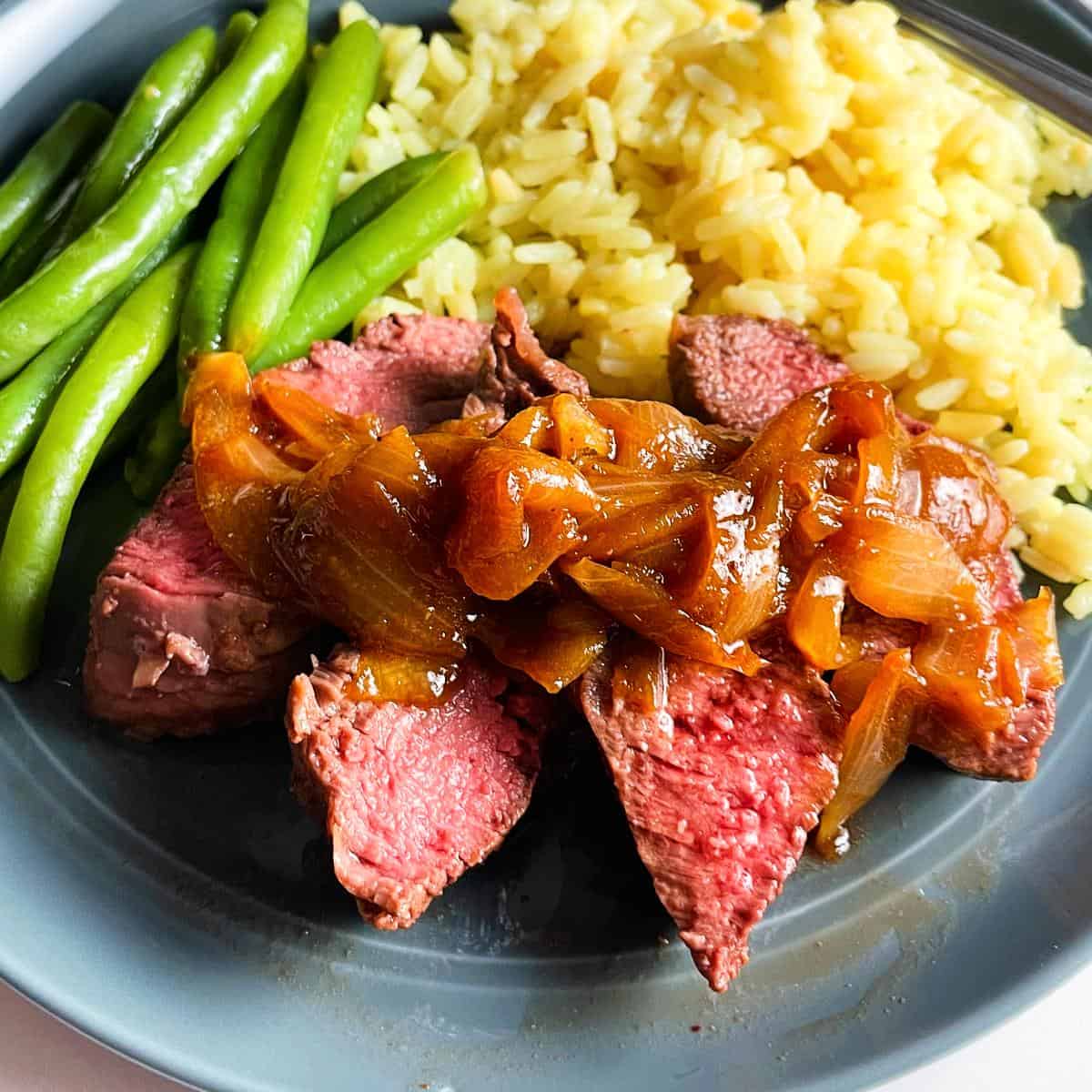 slices of oven roasted sirloin steak with onion sauce, on a plate with rice and green beans.