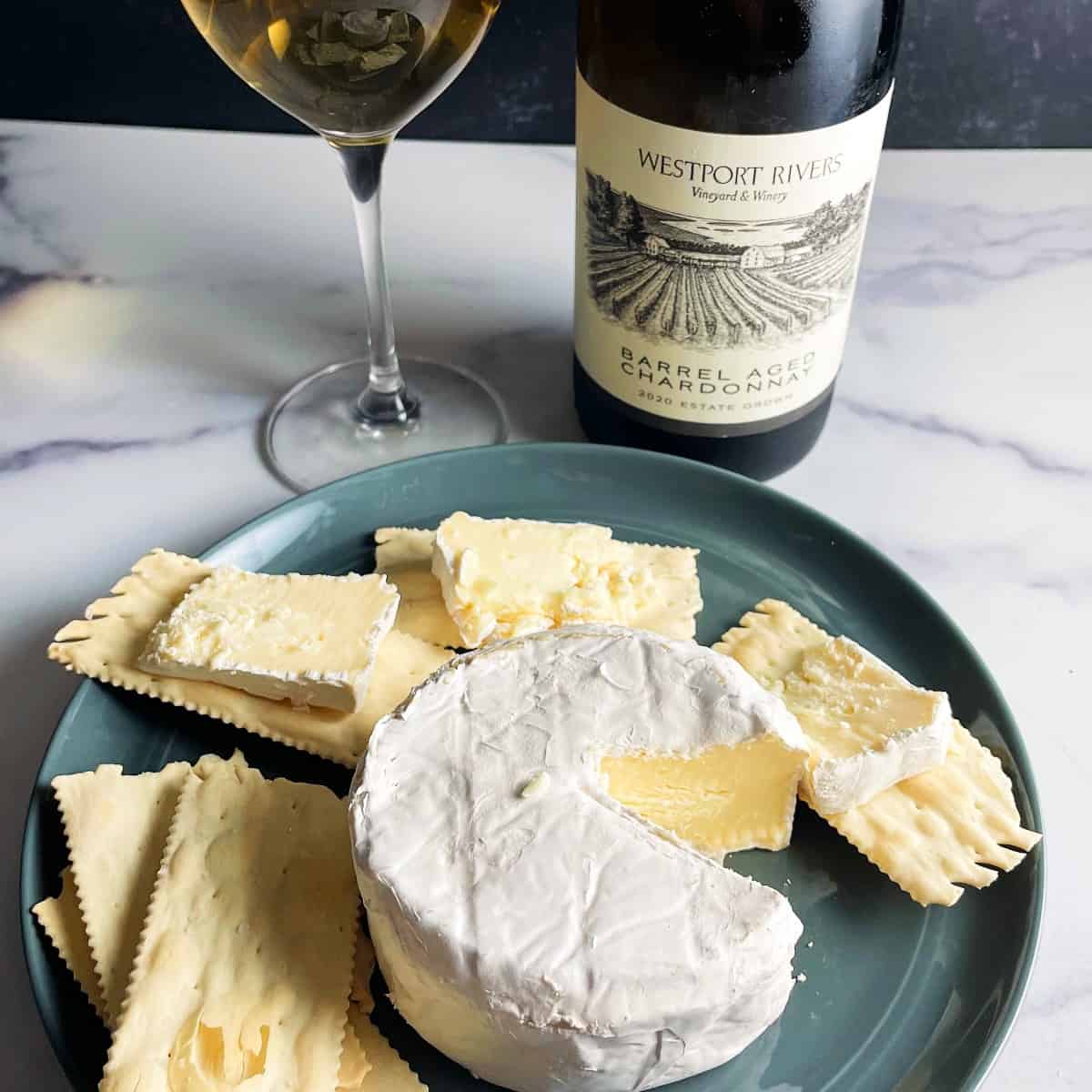 a wheel of brie on a plate, with a portion cut out and served on crackers. white wine in the background.
