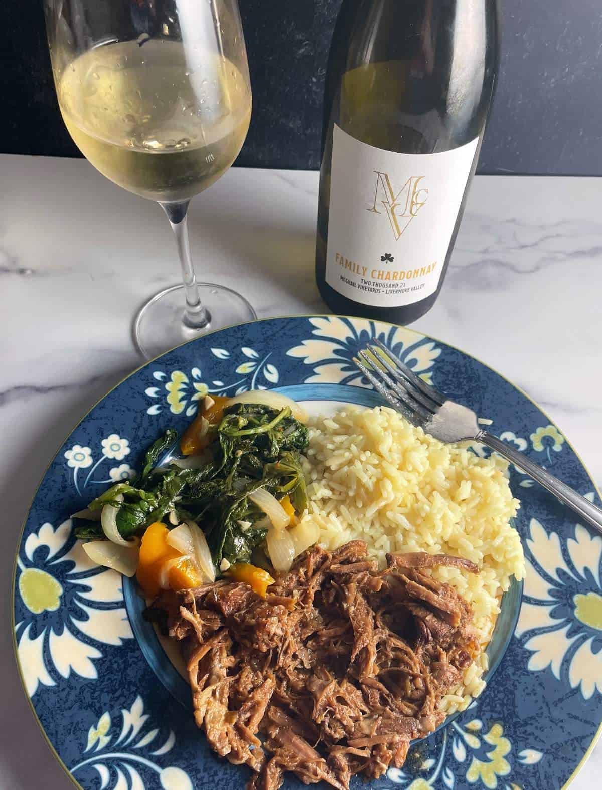 a plate with pulled pork, rice and sautéed greens, with a bottle and glass of Chardonnay white wine in the background.