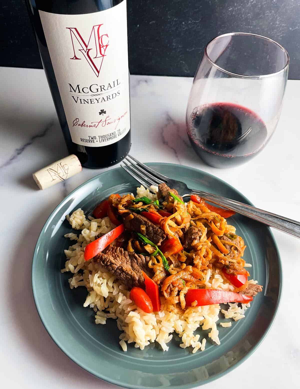 Stir-fried slices of steak and red bell pepper served over brown rice on a gray plate. A bottle and glass of Cabernet Sauvignon red wine in the background.