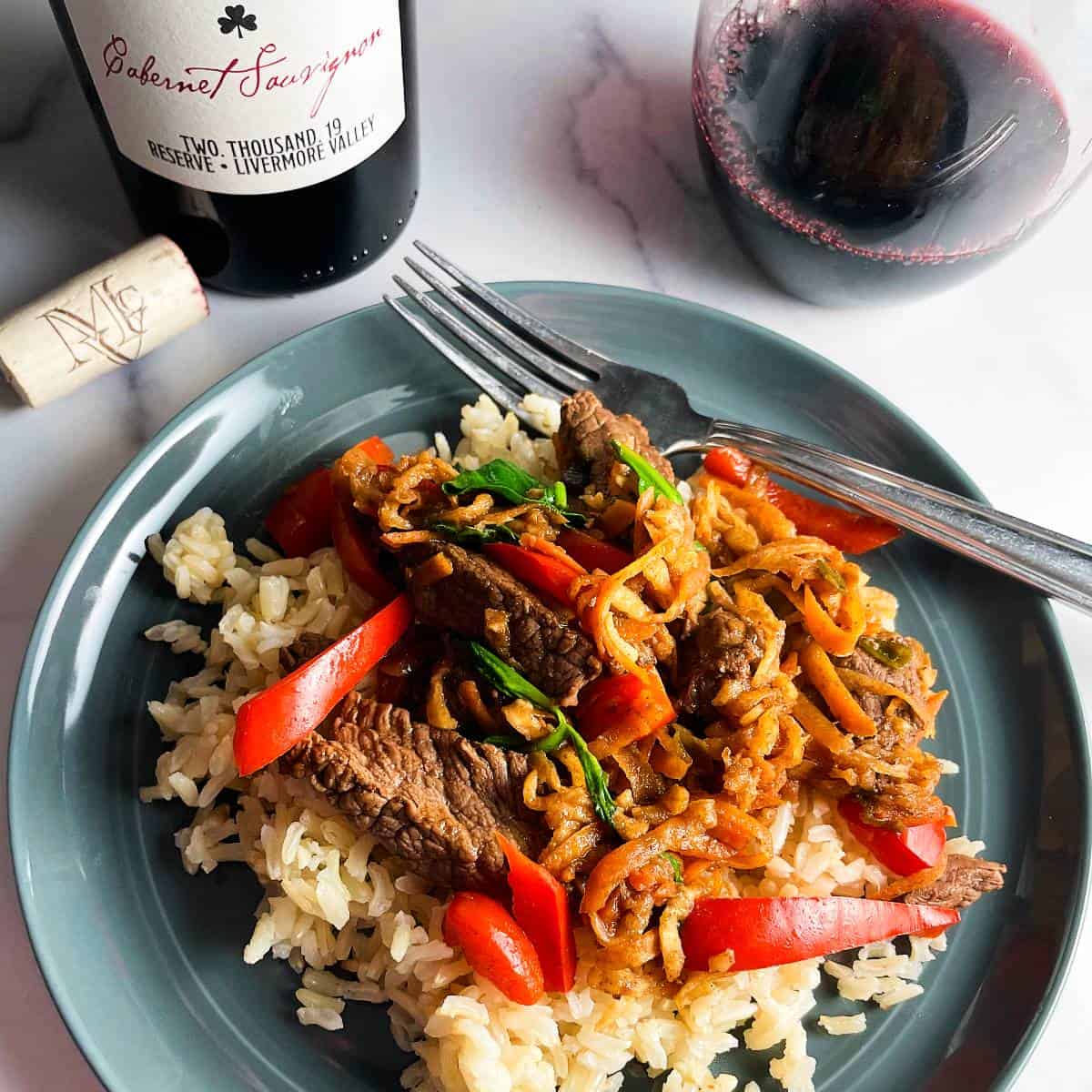 steak and vegetable stir-fry, including slices of red bell pepper, on a gray plate. Served with a red wine.