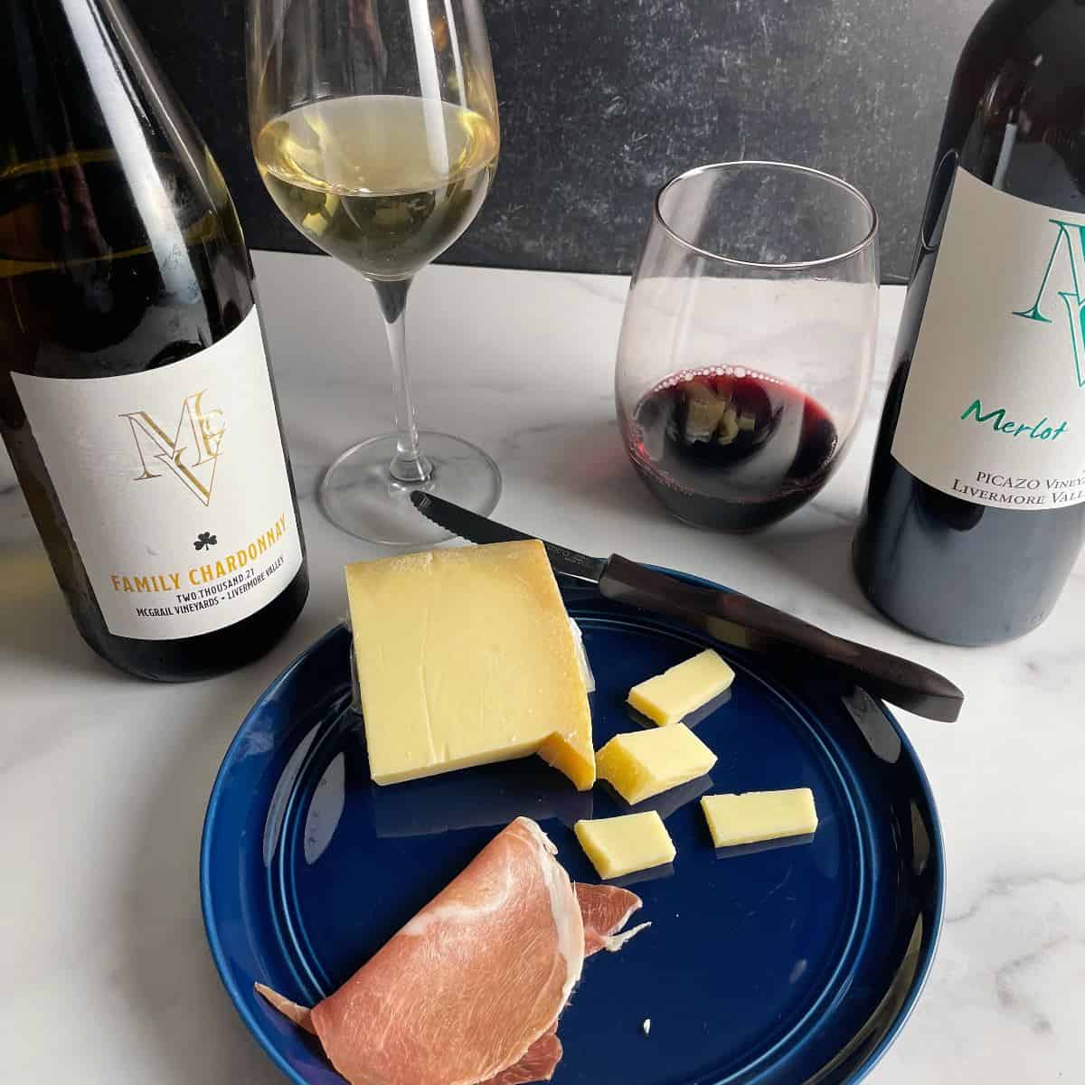 blue plate with Gruyere cheese and prosciutto, served with a red and white wine in the background.