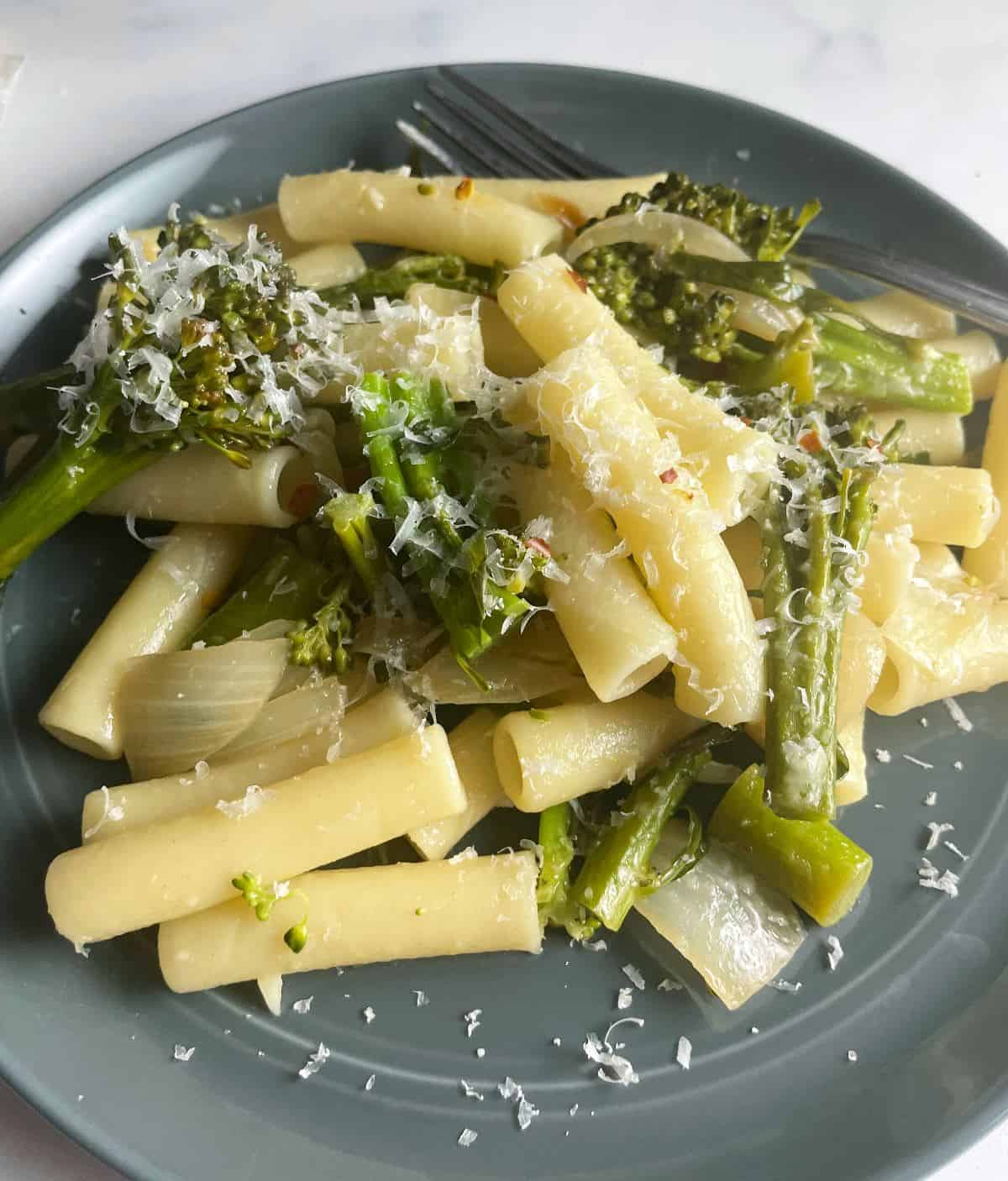 Broccolini tossed with ziti and shredded Parmesan cheese, on a dark gray dish.