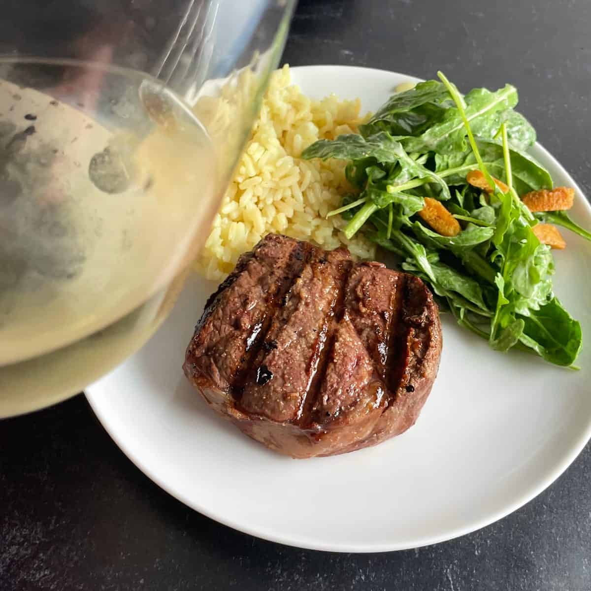 grilled filet mignon on a white plate with green salad and rice. A tilted glass of white wine off to the side.