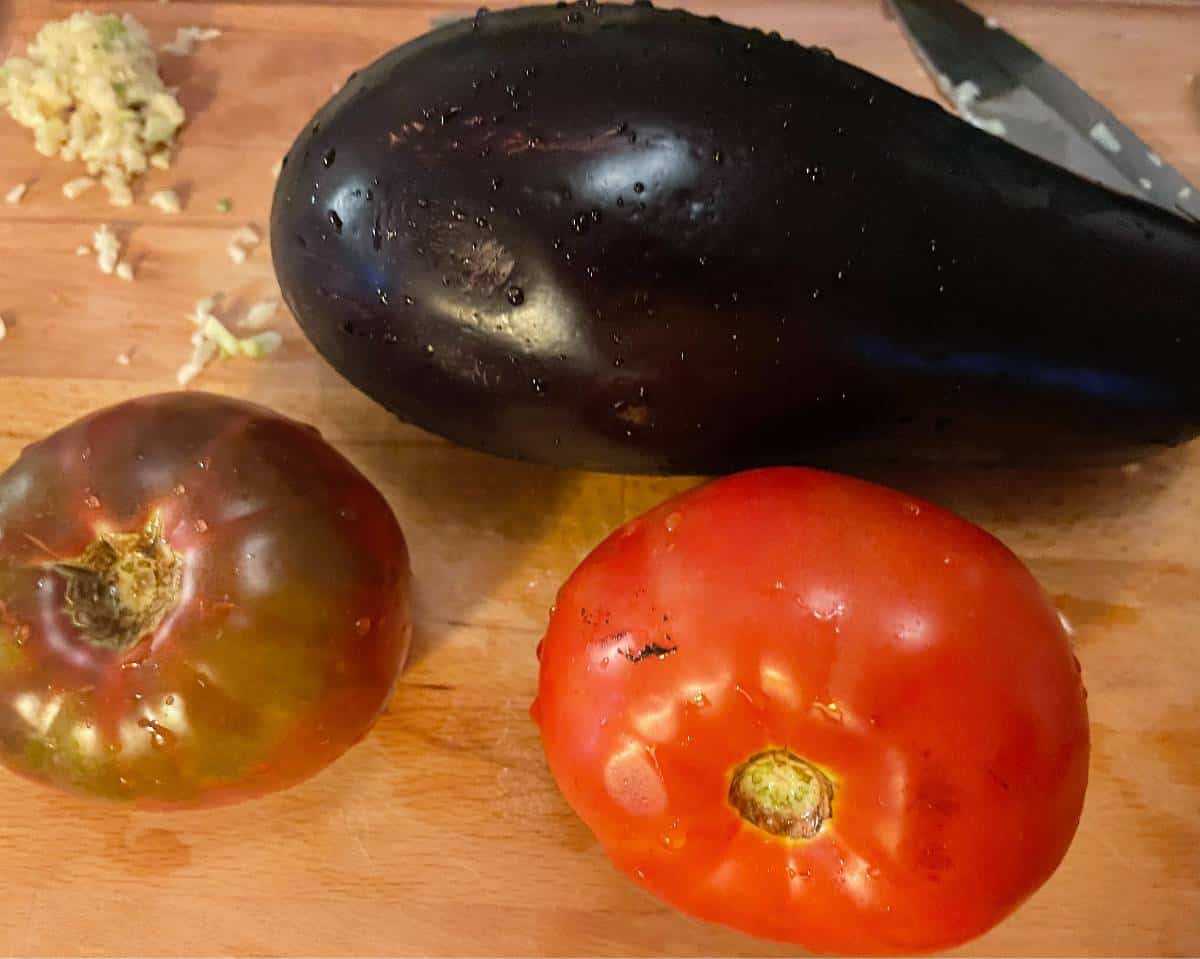 two large tomatoes and an eggplant on a cutting board, with some minced garlic on the side.