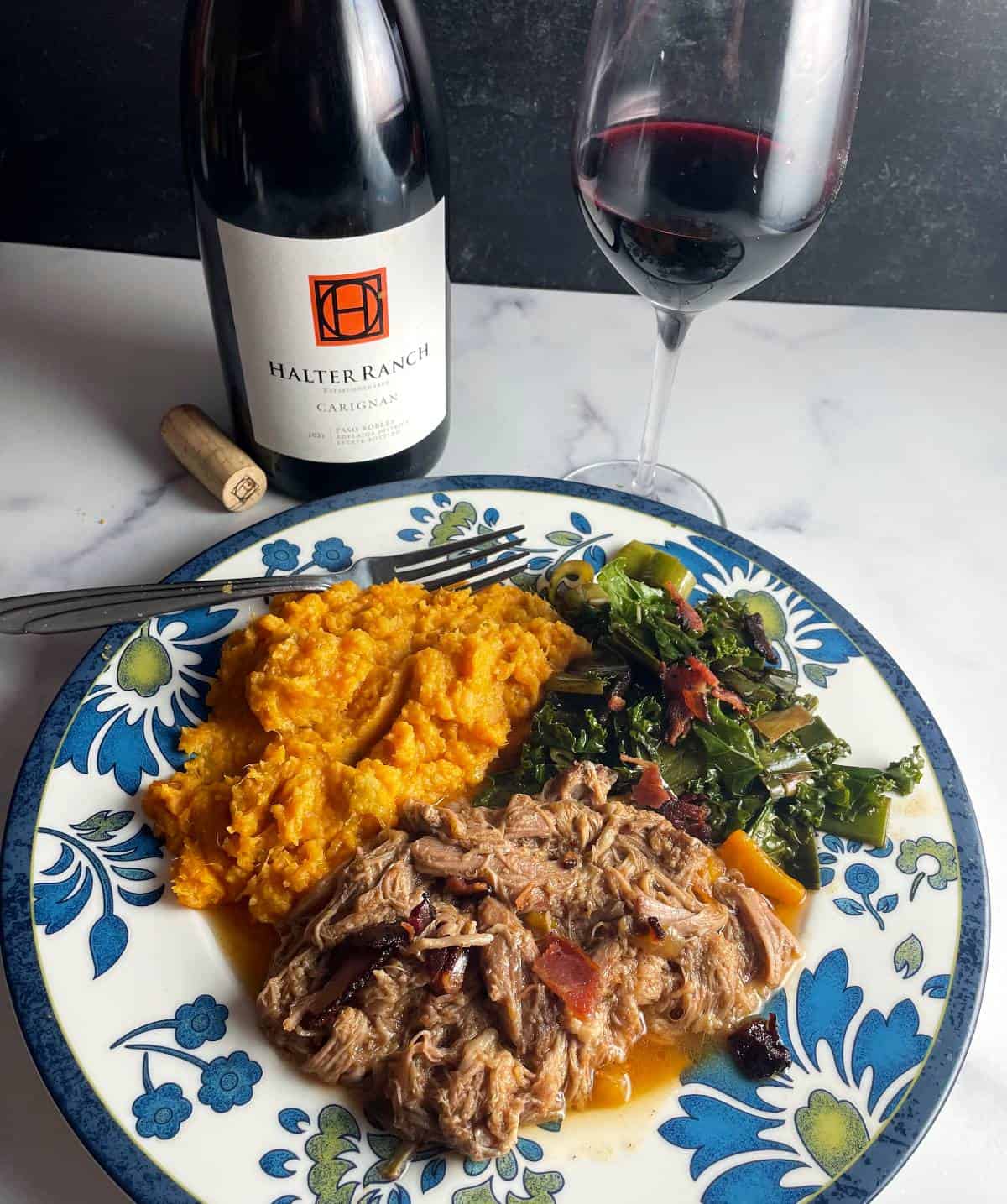 plate with flowers on the edges, with pulled pork, mashed sweet potatoes and leek greens. Bottle and glass of Carignan red wine in the background.
