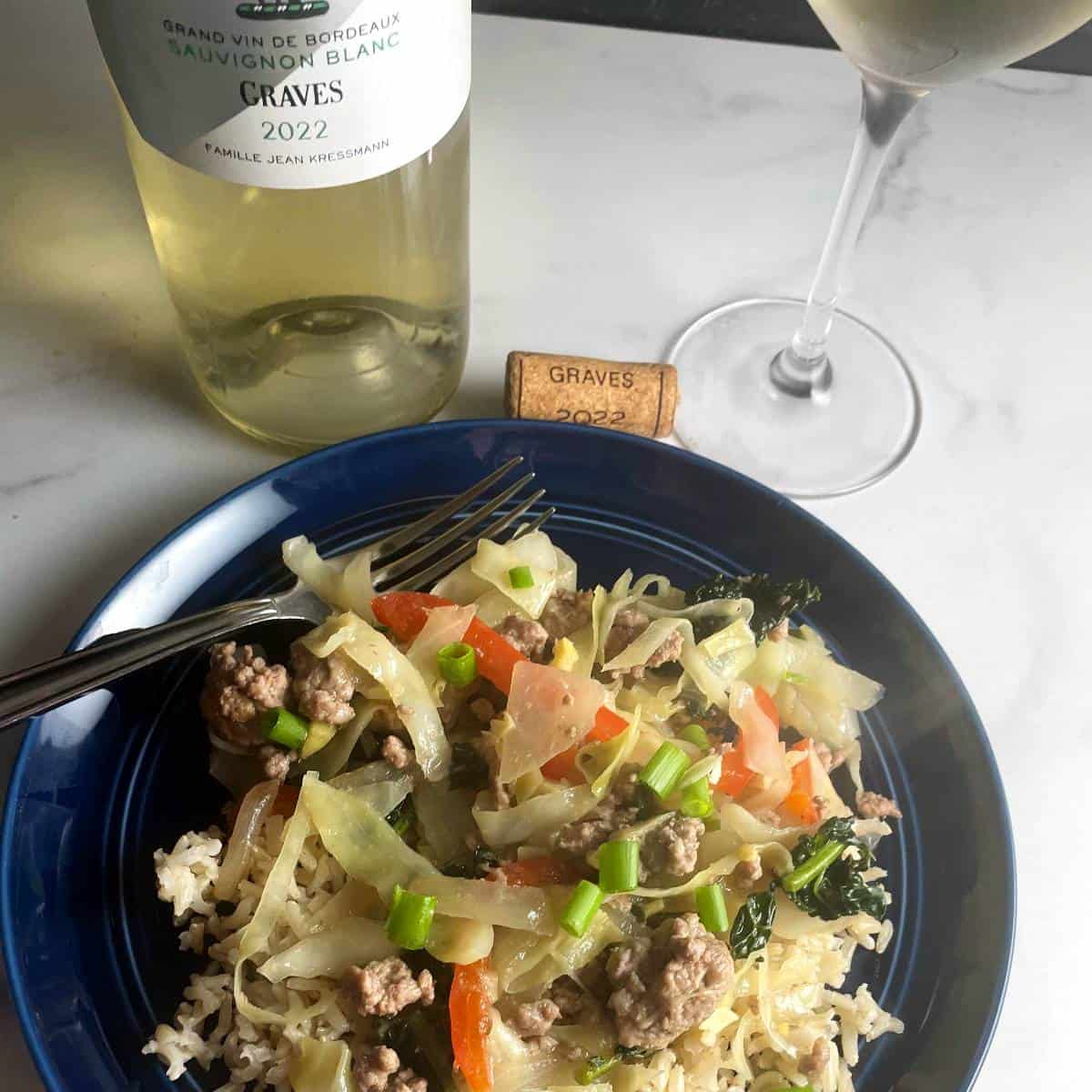 ground pork and cabbage stir-fry on a blue plate, served with white wine in the background.