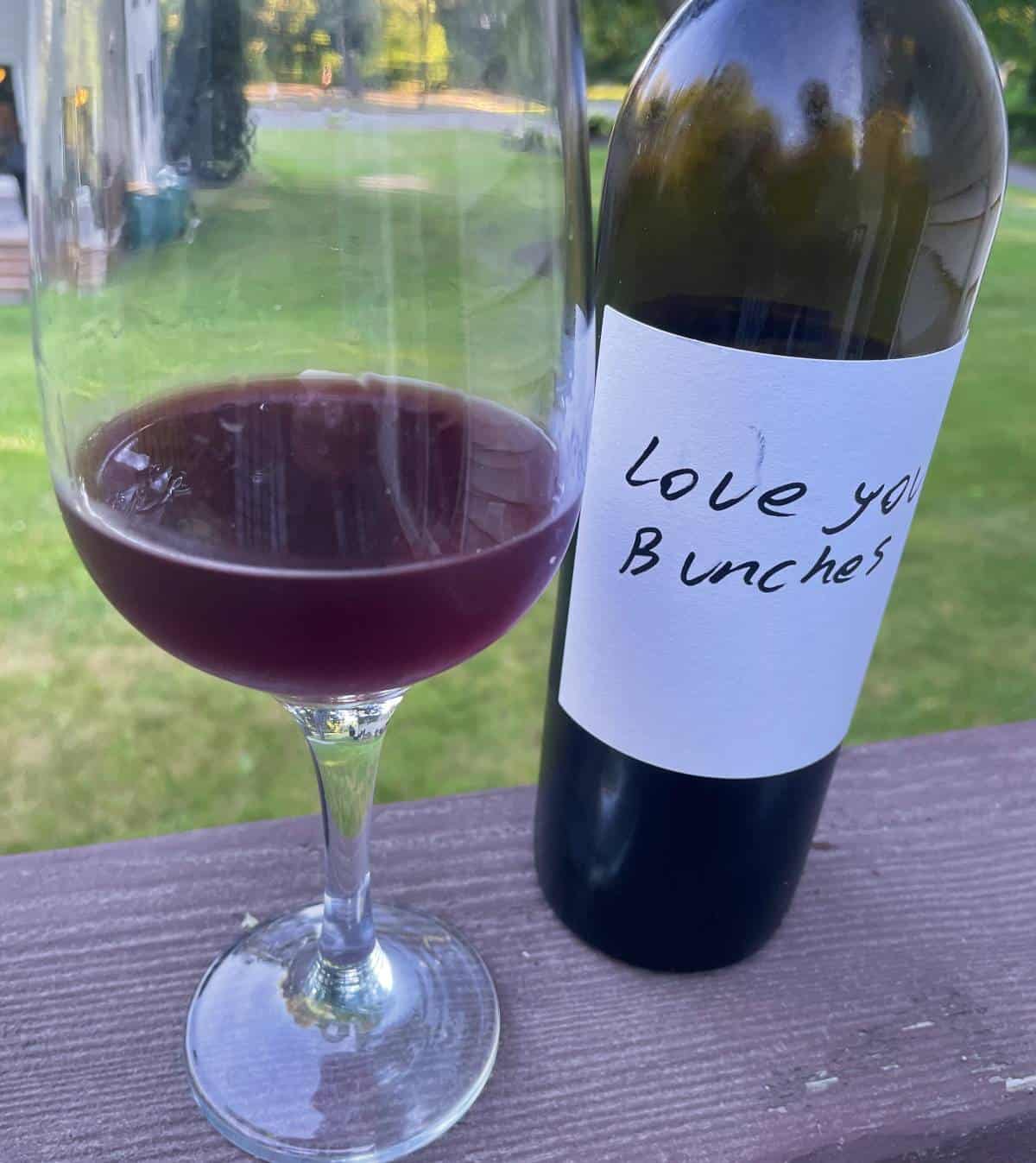 a bottle and glass of Love You Bunches red wine on a deck railing, with green grass in the background.