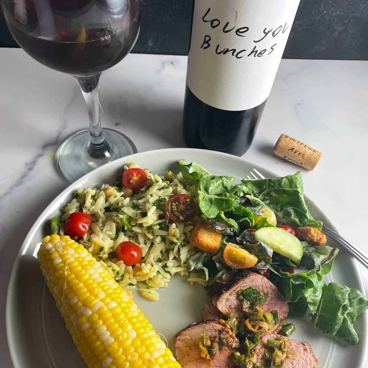 a plate with chard salad, pork tenderloin, corn on the cob, and orzo salad with kale pesto. A bottle of red wine and glass of it in the background.
