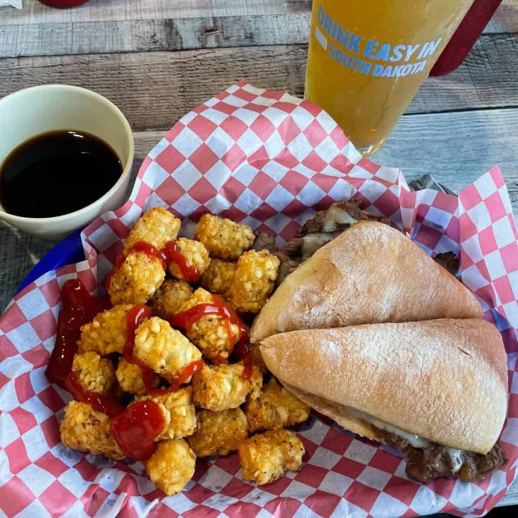 a roast beef dip sandwich with tater tots, shot from above with a glass of beer in the background.