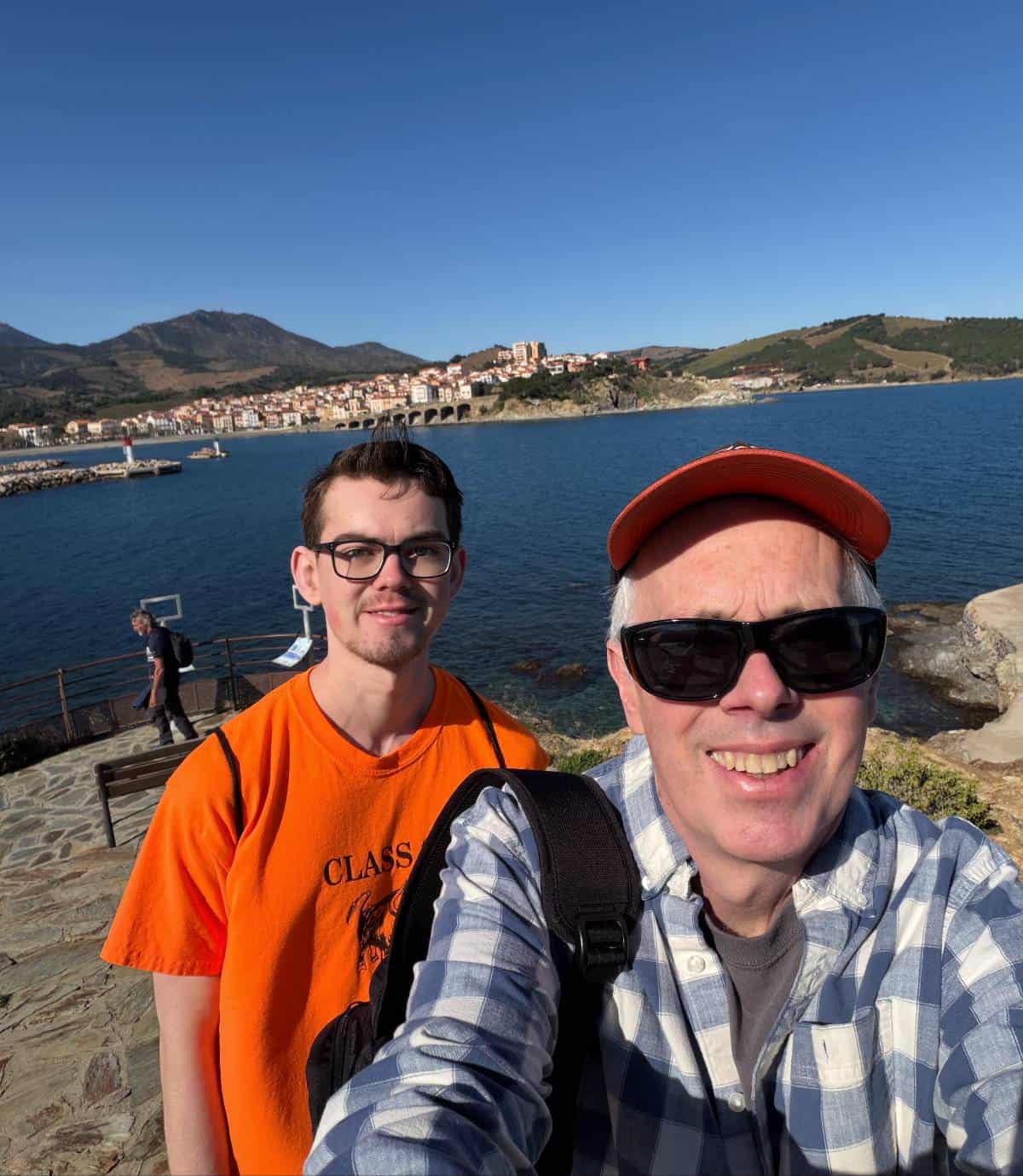 a father and son selfie pose with the Banyuls harbor behind them.