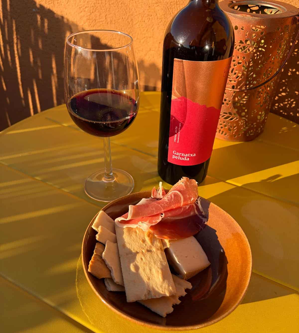 a bottle of red wine, with a glass poured, served on an outdoor table with cheese, crackers and jamon.
