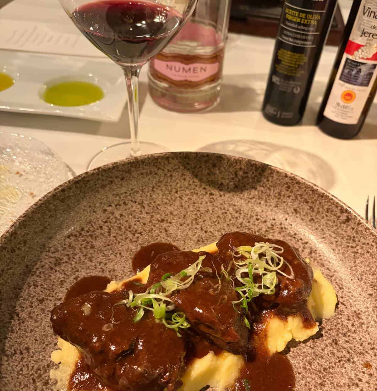 a plate with braised beef over potatoes and wine in the background.